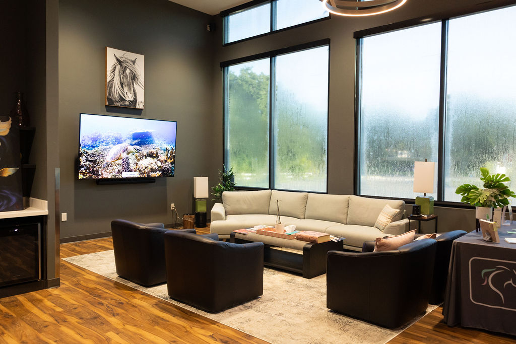 Modern waiting lounge with sectional sofa, dark chairs, TV, large windows, and hardwood floors